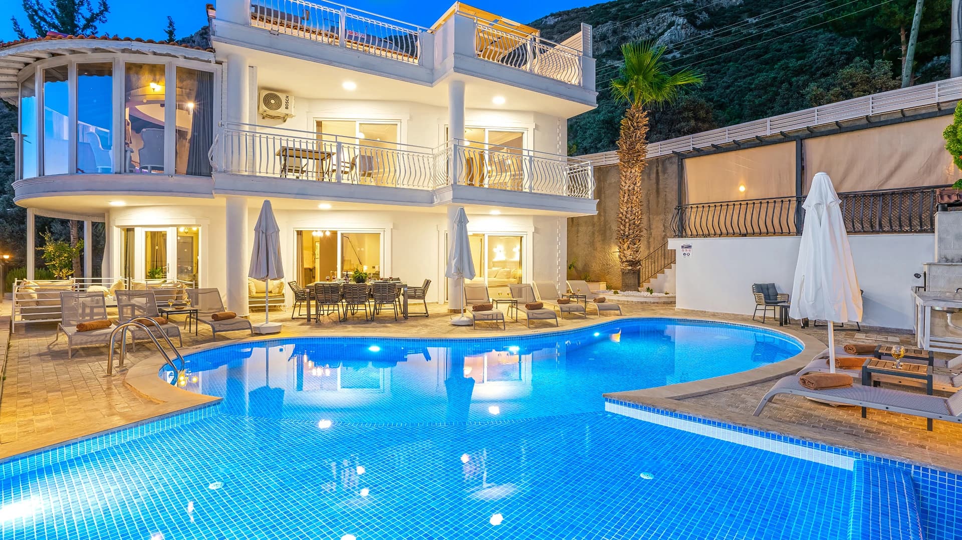 Illuminated private pool with expansive terrace, loungers, and mountain backdrop at dusk.