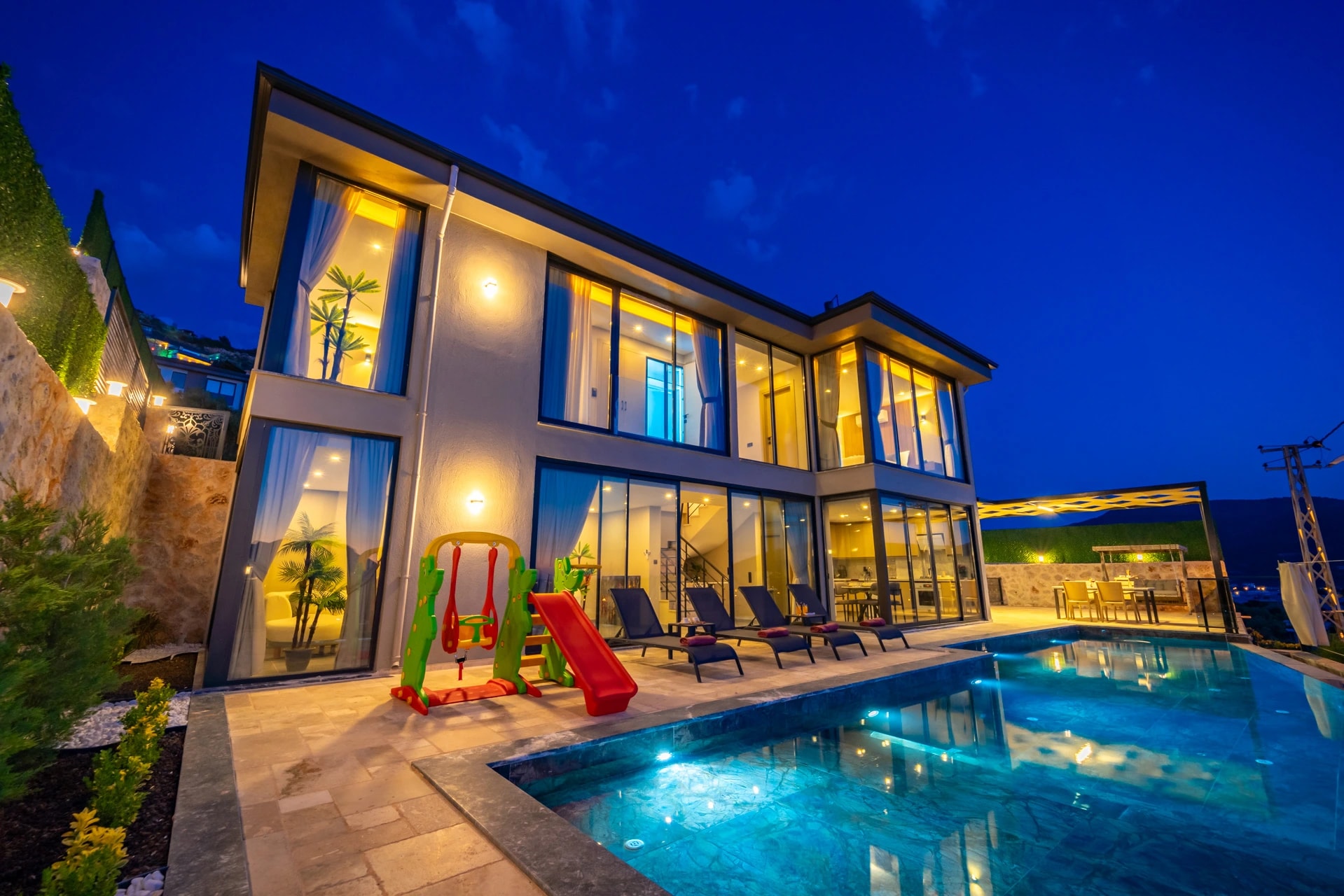 Modern villa exterior at twilight featuring heated pool, lounge area, and children's play structure.
