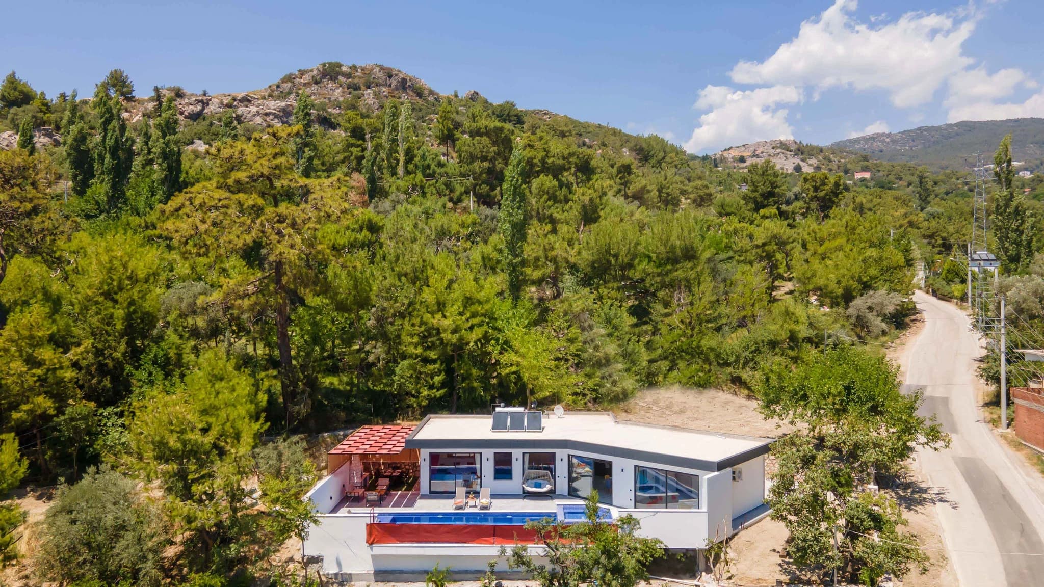 Kalkan'da Doğa Manzaralı, Özel Havuzlu ve Korunaklı Bahçeli Villa – Contemporary villa exterior nestled in hillside forest setting with pool, red terrace canopy, and modern architecture.