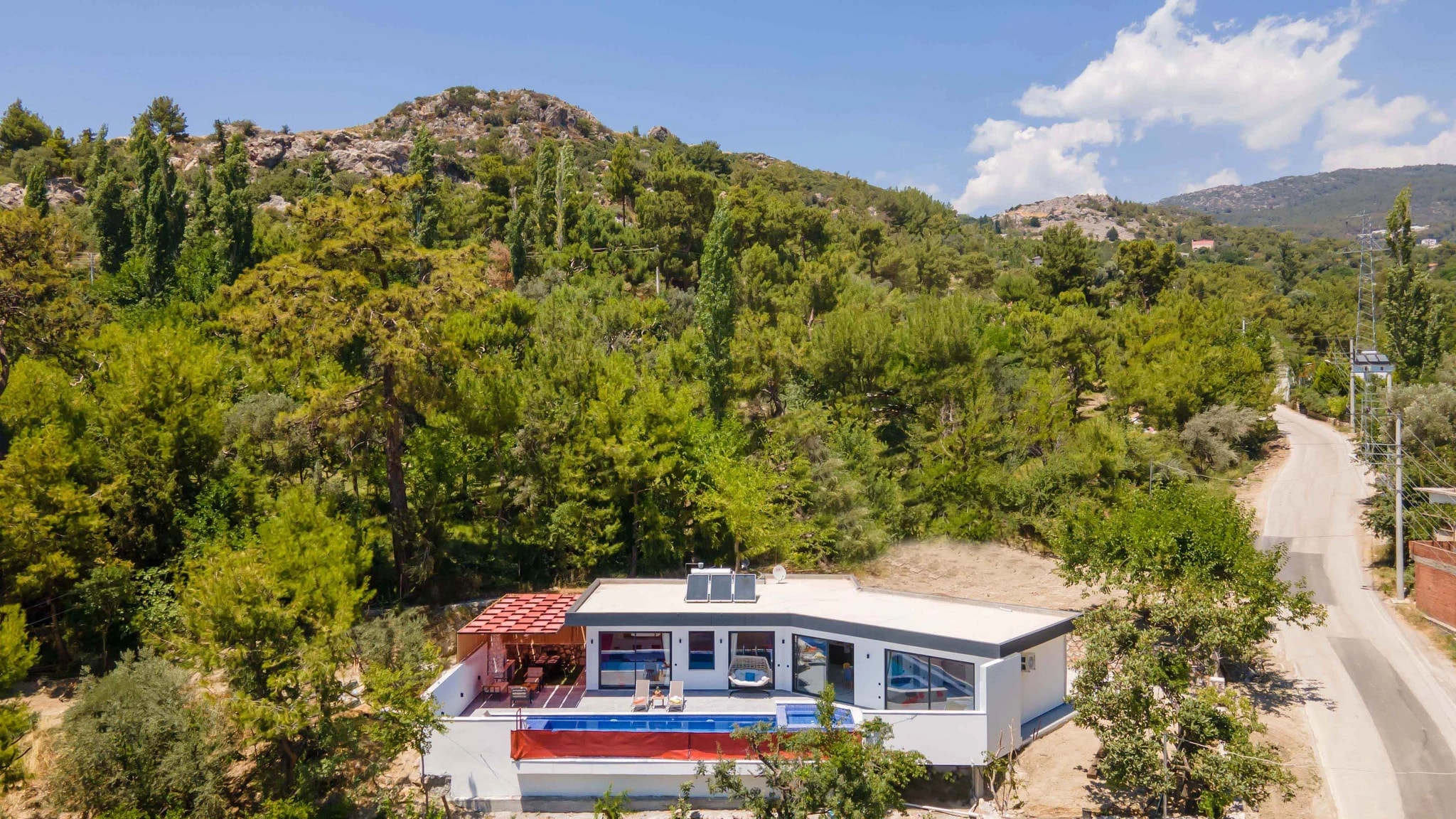 Kalkan'da Doğa Manzaralı, Özel Havuzlu ve Korunaklı Bahçeli Villa – Contemporary villa exterior nestled in hillside forest setting with pool, red terrace canopy, and modern architecture.