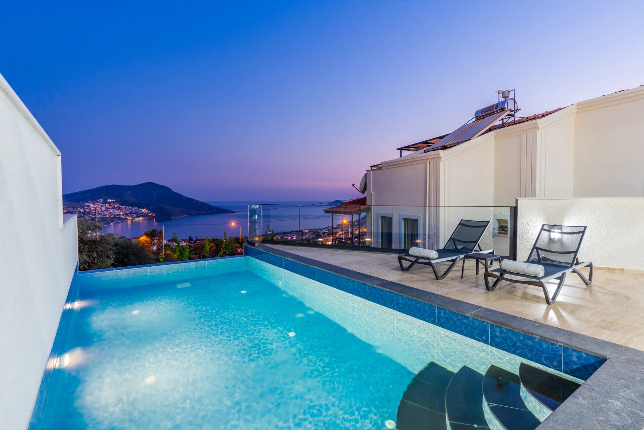 Elegant pool area overlooking illuminated coastal town and mountains at twilight.