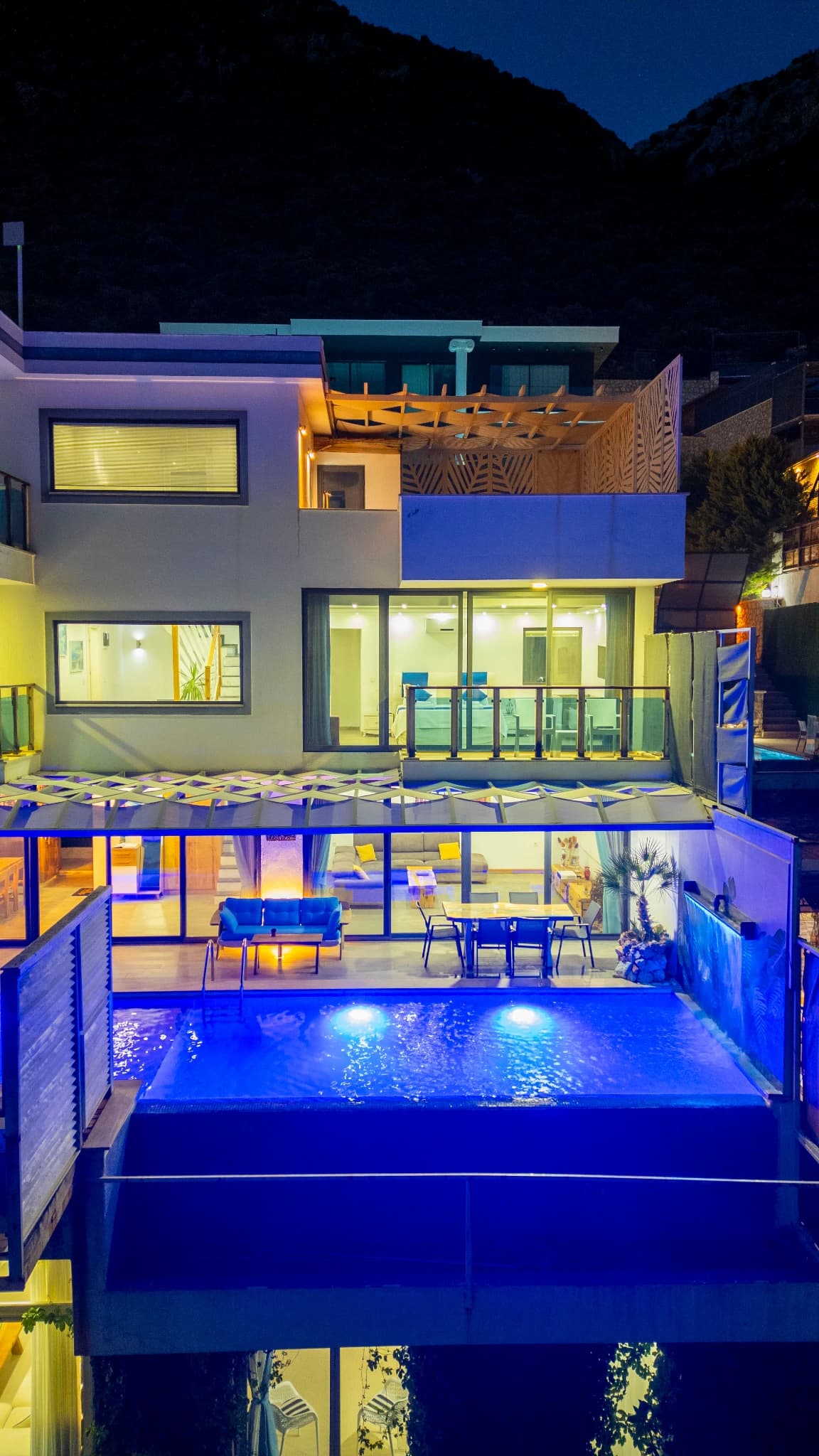 Evening view of illuminated modern villa featuring pools and terraces against mountain scenery.
