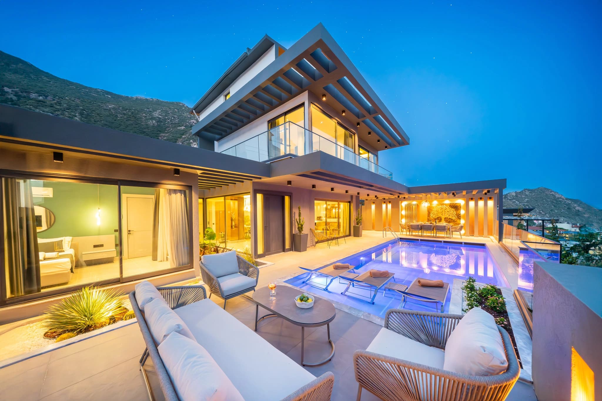 Illuminated private pool with loungers overlooking mountains at dusk.