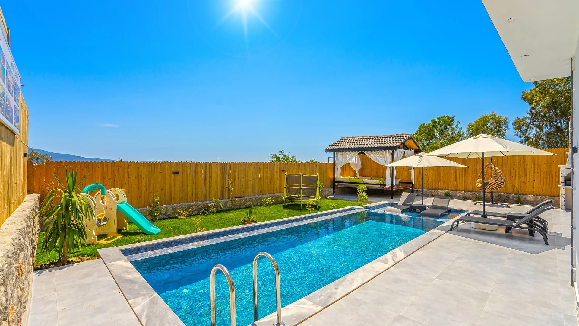 Pristine pool with sun loungers, parasols, and children's trampoline in spacious garden.