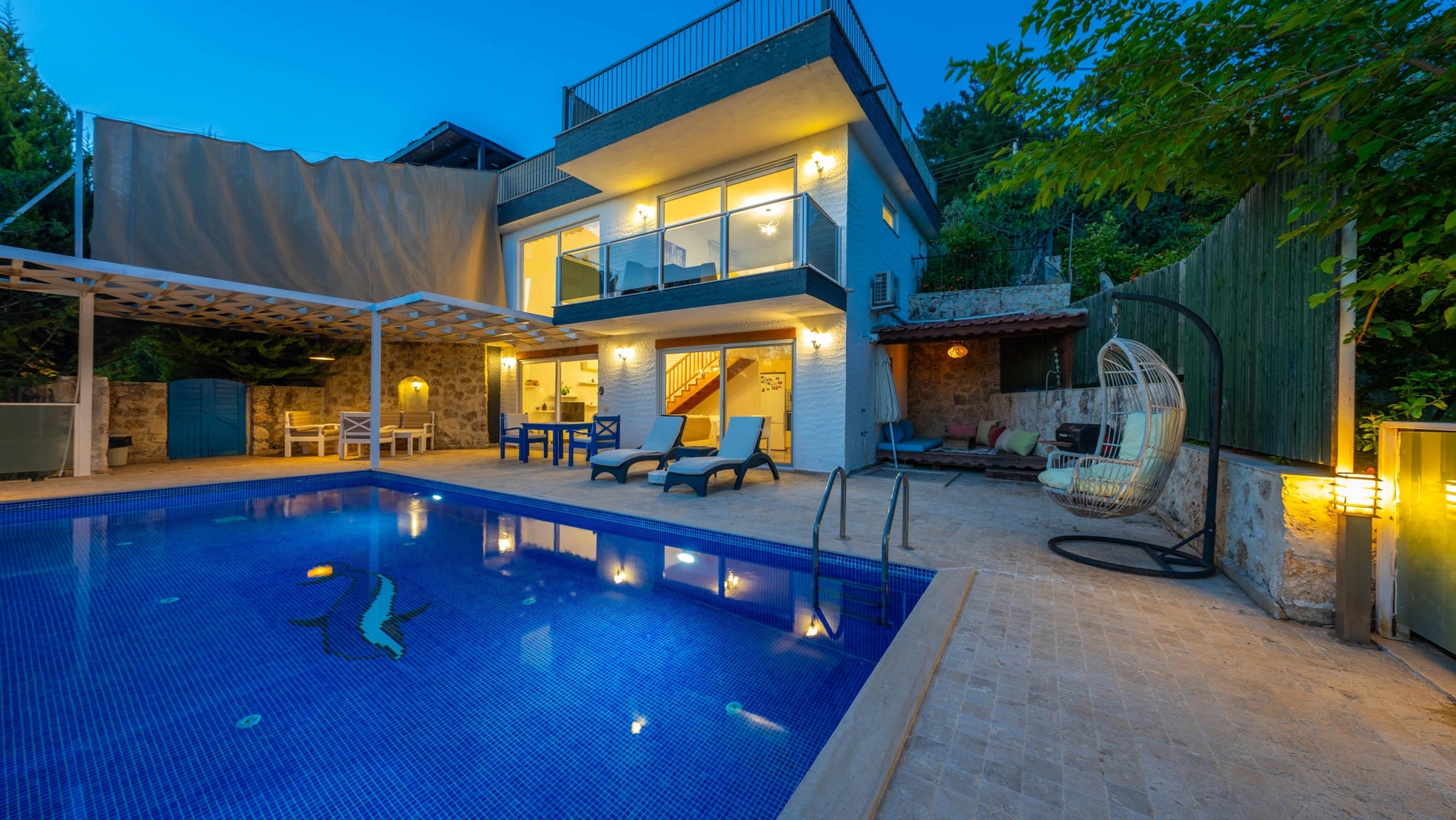 Evening view of illuminated private pool with lounge seating and pergola-shaded terrace.