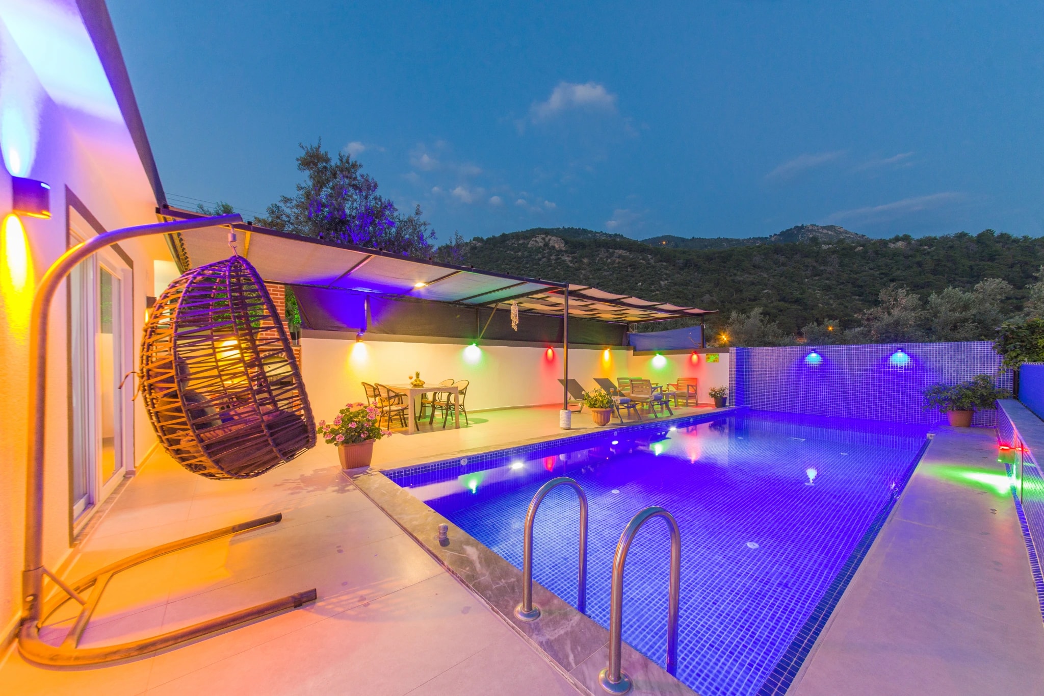 Pool with hanging chair, LED lighting, and forested hillside landscape in evening light.