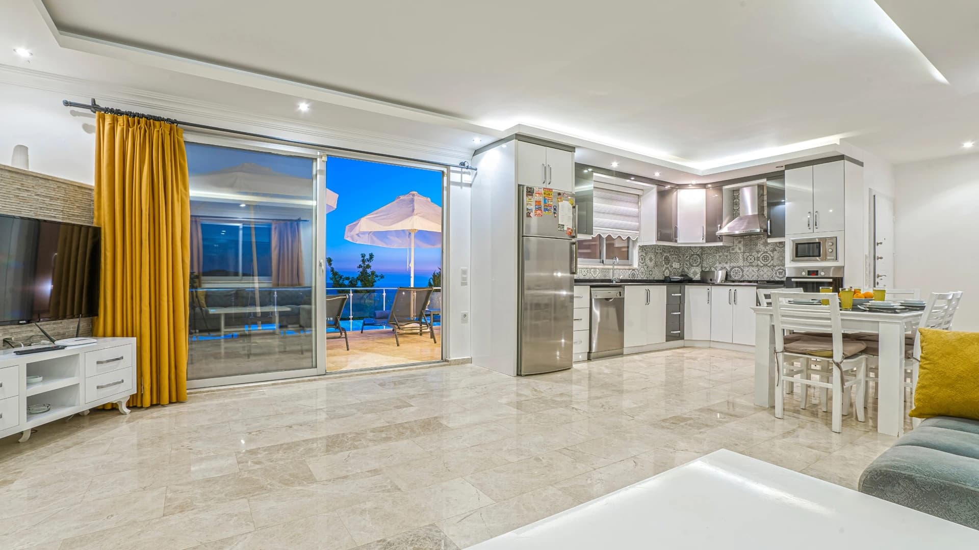 Kalkan Üzümlü'de Dağ ve Deniz Manzaralı, Isıtmalı Havuzlu, Korunaklı 2 Yatak Odalı Villa – Modern open-plan living and dining area with kitchen, overlooking pool and sea.