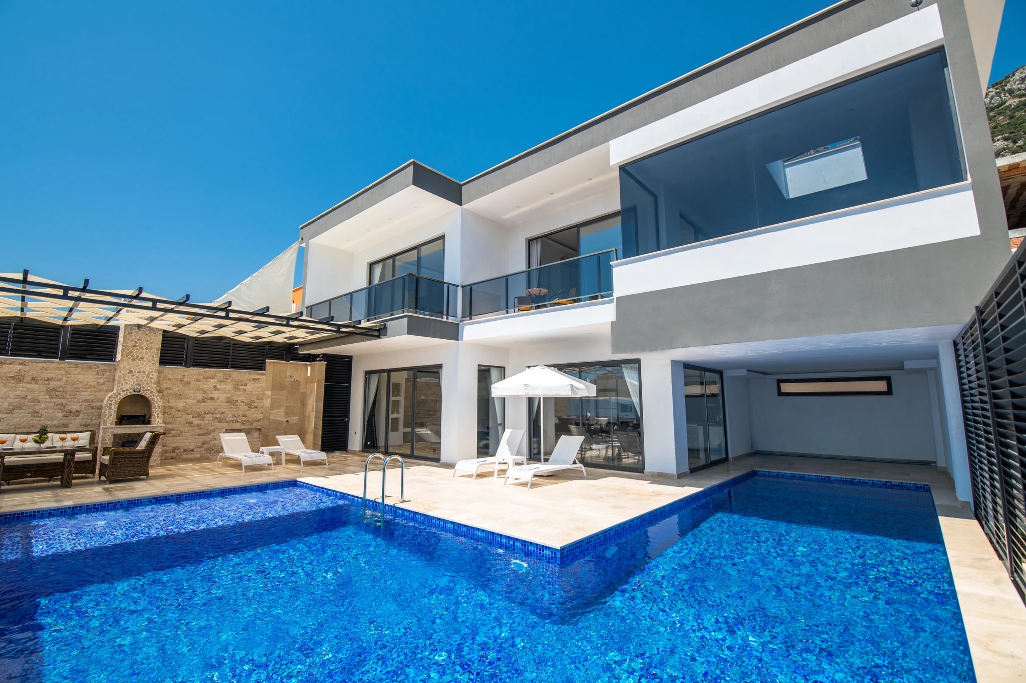 Kalkan'da Özel Havuzlu ve Isıtılabilen Kapalı Havuzlu Korunaklı Balayı Villası – Modern villa exterior with large swimming pool, sun loungers, and outdoor seating area in bright daylight.