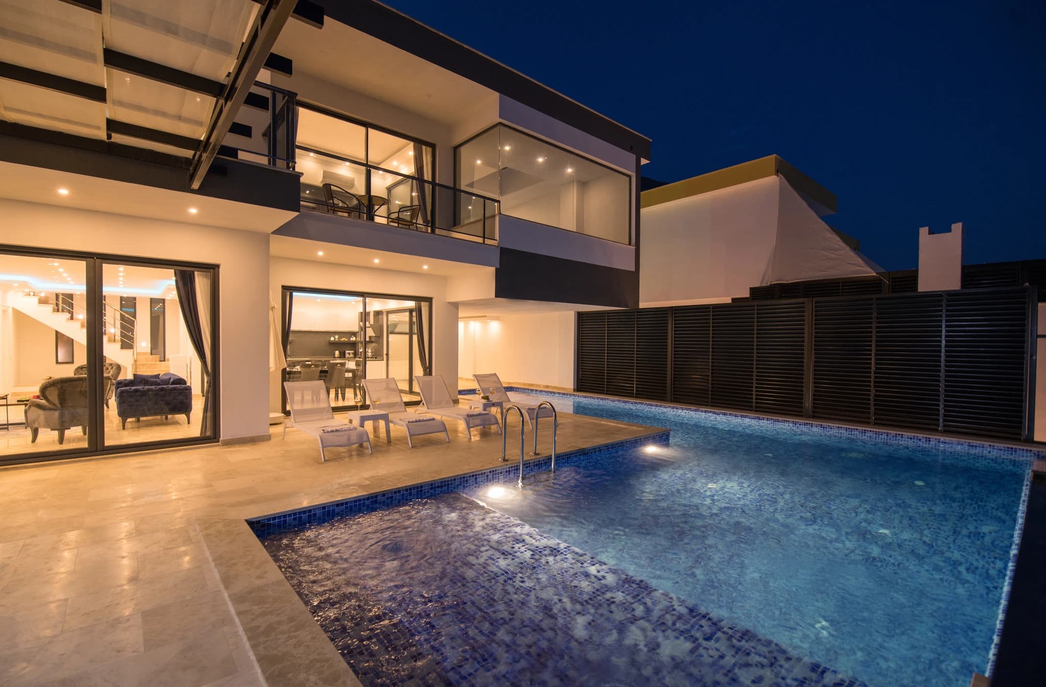 Kalkan'da Özel Havuzlu ve Isıtılabilen Kapalı Havuzlu Korunaklı Balayı Villası – Stunning nighttime pool area with illuminated water, sun loungers, and modern villa interior visible through glass doors.