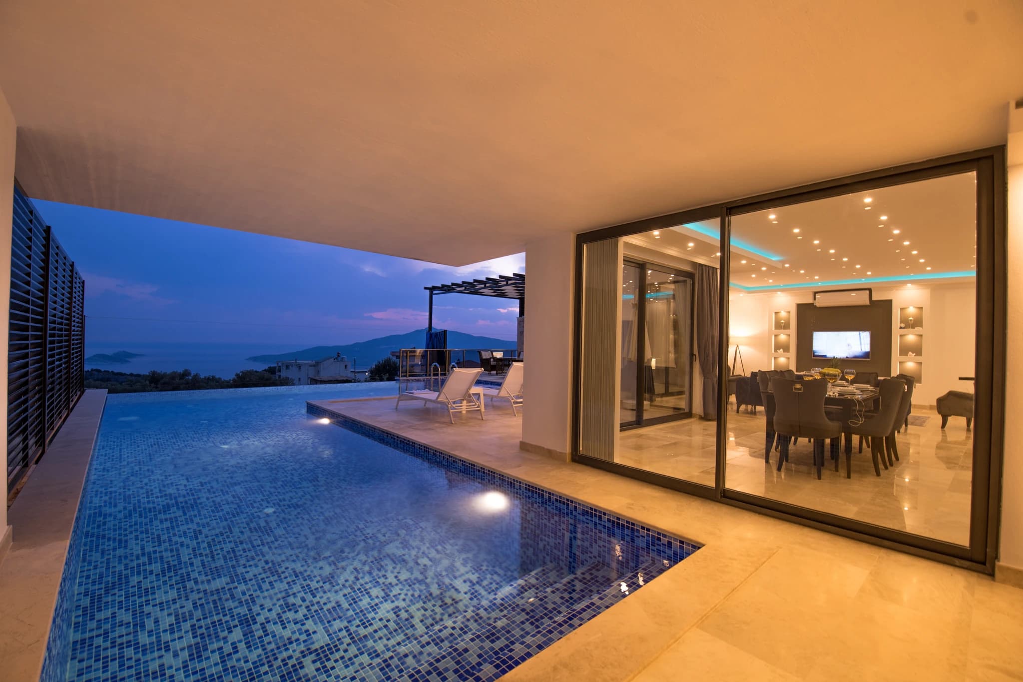 Kalkan'da Özel Havuzlu ve Isıtılabilen Kapalı Havuzlu Korunaklı Balayı Villası – Stunning illuminated pool overlooking mountains, with adjacent terrace and lounge chairs.