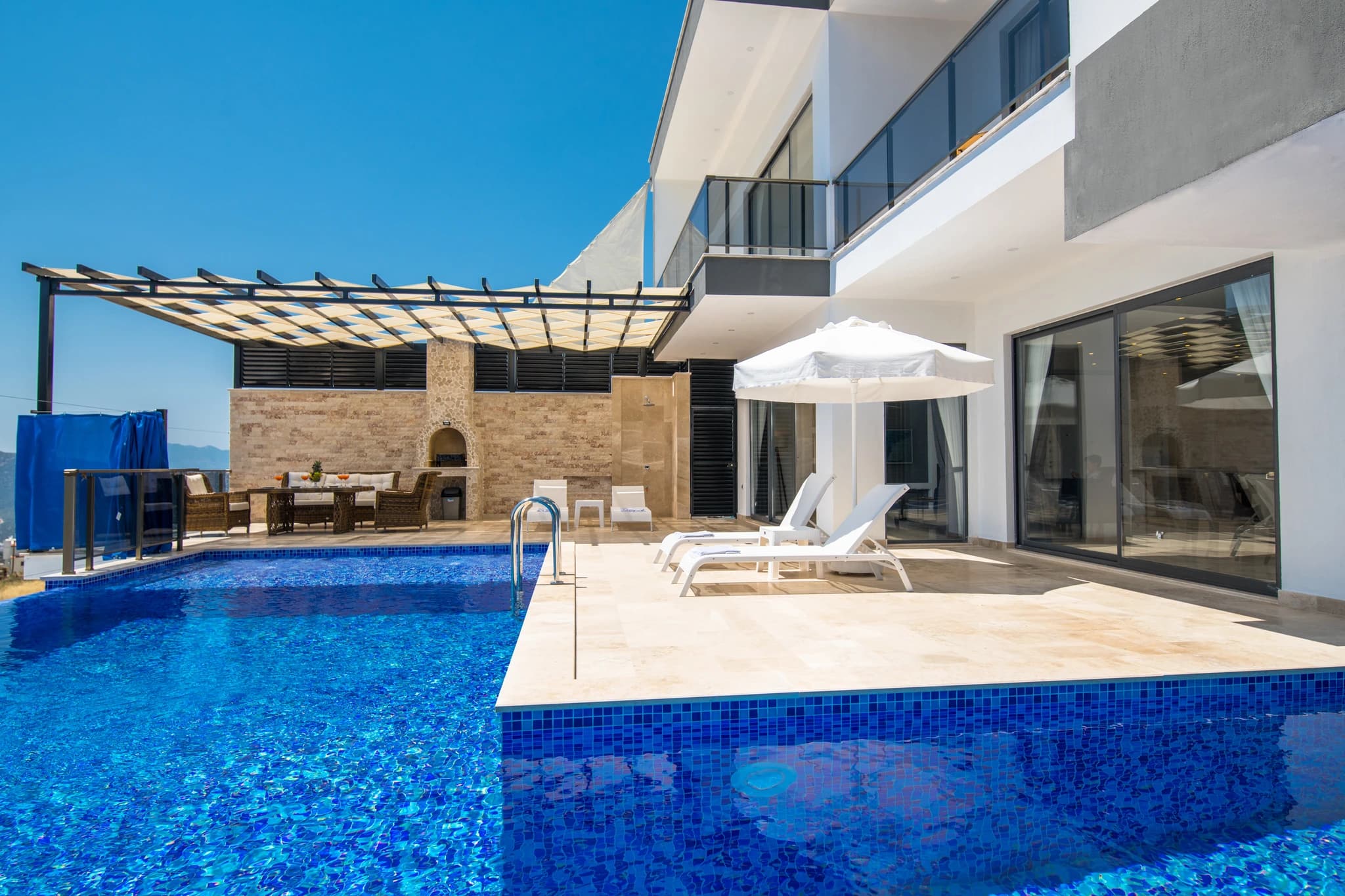 Kalkan'da Özel Havuzlu ve Isıtılabilen Kapalı Havuzlu Korunaklı Balayı Villası – Resort-style pool area with dual pools, lounge chairs, pergola shade structure, and stone fireplace on expansive terrace.